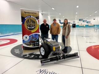 Three community representatives beside sponsored ice-maintenance equipment at a curling rink.